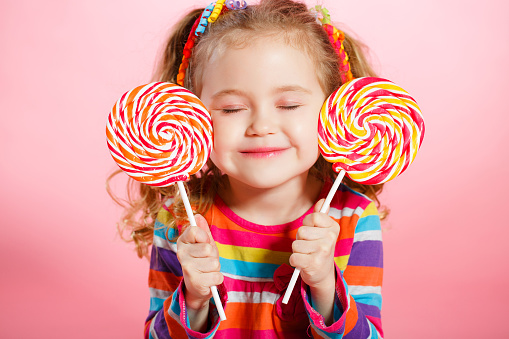 Funny little girl with long, curly red hair,bright ribbons tied into two tails, a sweet smile,wearing a bright dress with a red bow on the chest,posing in Studio on pink background holding two big colorful Lollipop