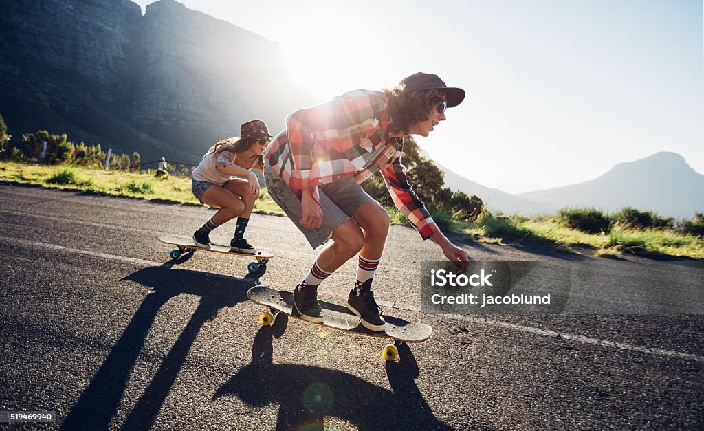 Young friends longboarding down the road Side portrait of young people skateboarding together on road. Young man and woman longboarding down the road on a sunny day. Skateboarding Stock Photo