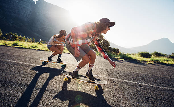 Young friends longboarding down the road Side portrait of young people skateboarding together on road. Young man and woman longboarding down the road on a sunny day. longboarding stock pictures, royalty-free photos & images