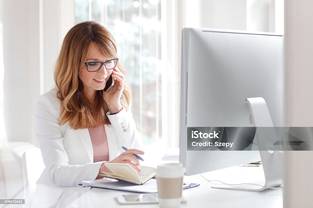 Day planning Portrait of an attractive businesswoman sitting in front of computer and taking notes while speaking on her cellphone 40-49 Years Stock Photo