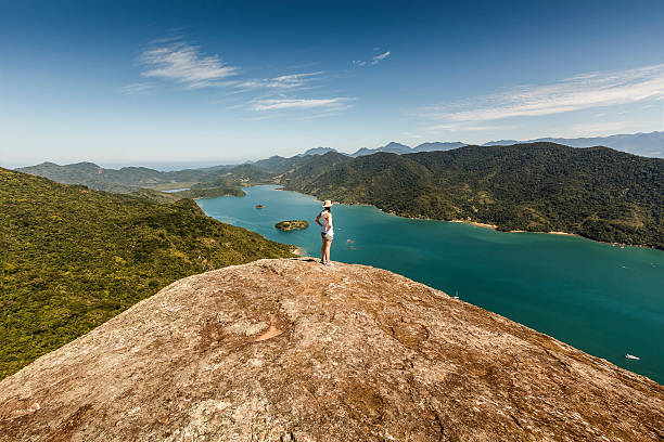 Young Woman on Top of High Mountain on Sunny Morning A DSRL photo taken at the top of a mountain beside the calm sea in Mamangua, Paraty, Rio de Janeiro, Brazil. At the very top there is a young woman contemplating the view. It is a sunny day with bright blue sky.  eco tourism photos stock pictures, royalty-free photos & images