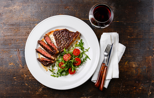 Sliced medium rare grilled Beef steak Striploin, salad with tomatoes and arugula on white plate and red wine on dark wooden background