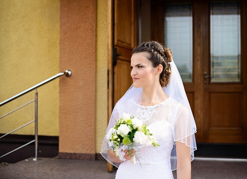 Happy young Bride standing in front of the church holding a bridal bouquet after the wedding ceremony. Sunny Wedding Day.