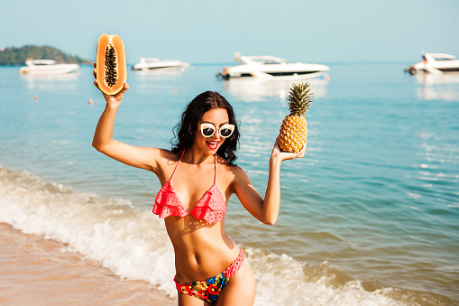 Pretty cool fashionable girl in the bright bathing suit, sunglasses, posing against the backdrop of the sea. In the hands holding a papaya and pineapple.