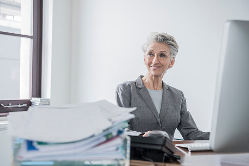 Mixed race mature businesswoman using computer. Happy woman in her 60s working in modern office, smiling, looking away from camera. Filing tray with paperwork in foreground.