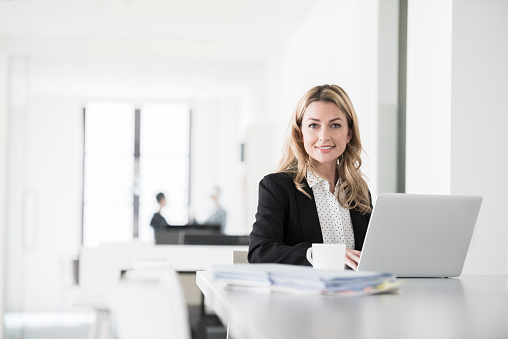 Candid portrait of mid adult businesswoman sitting at desk with laptop computer. Female office worker with long blonde hair smiling towards camera.