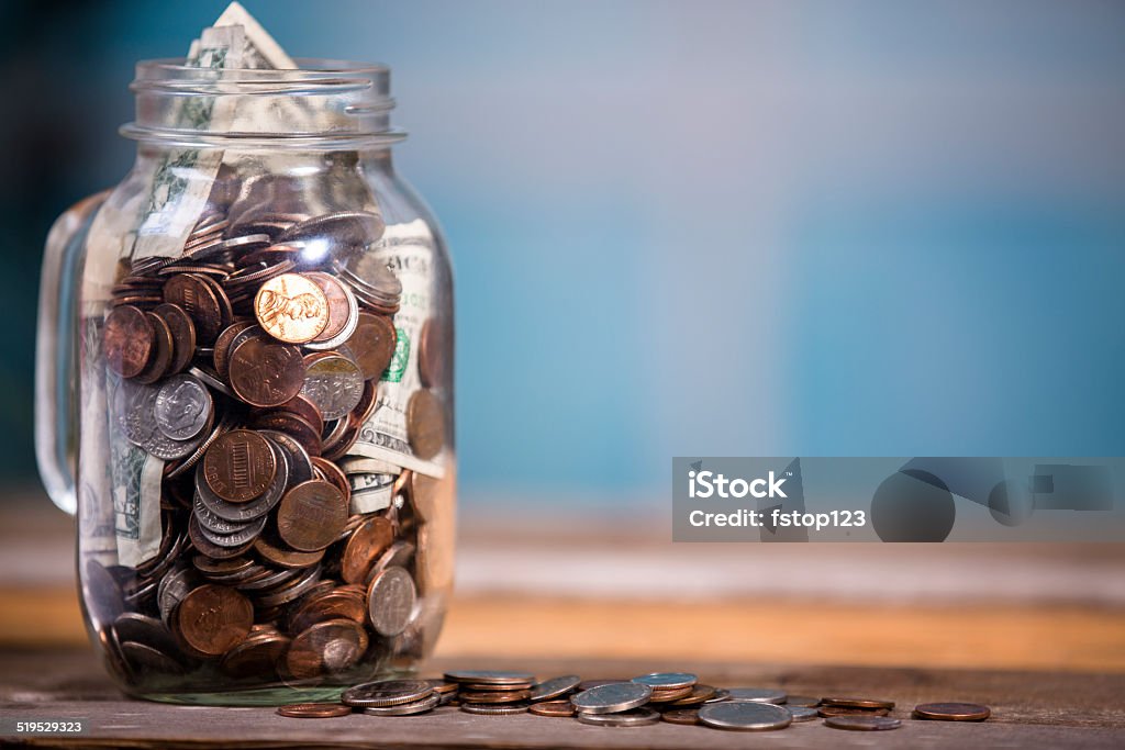 Money jar with U.S. currency.  Savings, donations concepts. Money Jar filled with US currency: coins and paper money.  Savings or donations concept. Blue window background.  American Culture Stock Photo