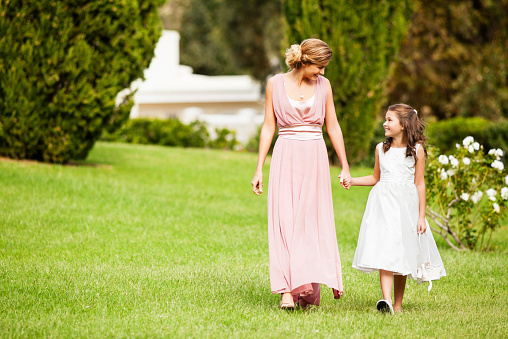 Happy bridesmaid and flower girl looking at each other while walking in garden. Horizontal shot.