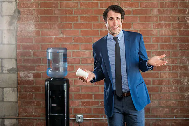 Obnoxious nerdy caucasian businessman mid thirties, wearing a blue suit.He is holding a coffee cup standing by the office water cooler.  Shot in the studio on a grey background with the Nikon D800.