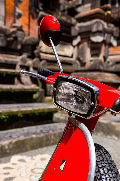 Ubud, Bali, Indonesia - August 13, 2014: Headlight and front of a red scooter with a Hindu temple in the background. The temple has oriental decorations; its dark colors contrast beautifully against the bright red color of the moped. The image evokes the thought of the adventurous journey and discovery in retro style.