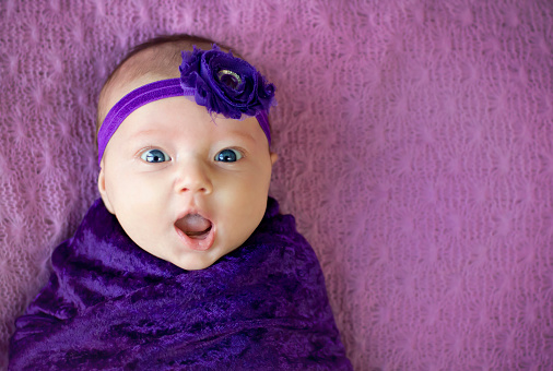 Happy and surprised expression on a 2 month old baby girl, with copy space. Purple background.