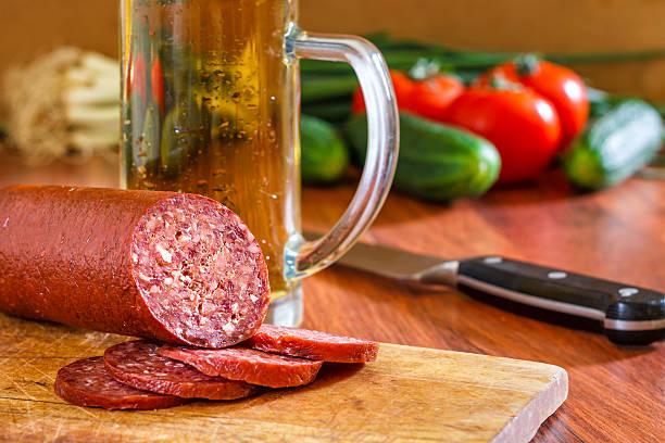 Sujuk sausage on wooden cutting board with beer mug, knife and fresh vegetables in background. Copy space