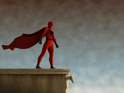 A businessman wearing a cape and a superhero mask about to leap a tall building with a big city skyline in the background. Standing on a cement rooftop, the tall building he faces looms overhead, yet he is not daunted.  With his red cape and black eye mask he stands ready to make the leap. 