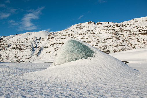 Blue iceberg on a frozen glacial lake at Skaftafellsjokull glacier at winter, on the background of rocks covered by snow, blue sky, Iceland