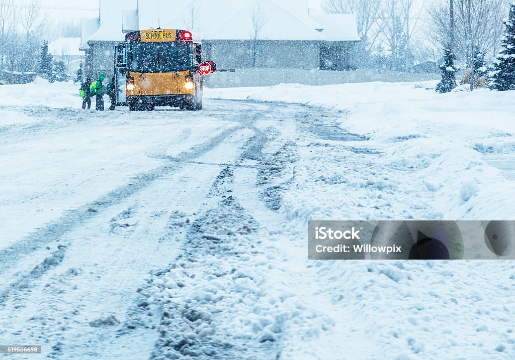 School Students Boarding Blizzard Snow Storm School Bus A line of anonymous school children students waiting to board a bright yellow school bus with open door, flashing red lights and outstretched warning stop sign during a raging winter blizzard snow storm on a slippery, slushy, tire track criss-crossed suburban residential street. School Bus Stock Photo