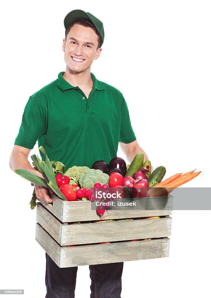 Delivery man with organic food Delivery man delivering box with organic food and smiling at the camera. Studio shot, white background. Supermarket Stock Photo