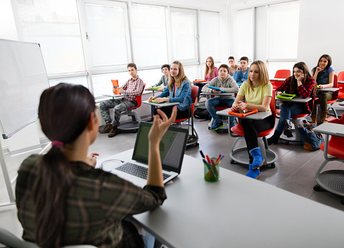 Large group of smiling students sitting in a modern classroom and listening to their teacher talking.