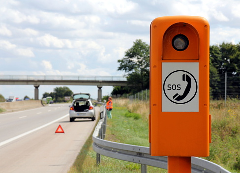 Emergency telephone on a German highway (Autobahn) with car broken down in the background, hazard flashers turned on, breakdown triangle in position and driver wearing reflective vest waiting behind crash barrier for breakdown service.