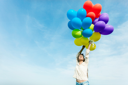 Young woman holding multi colored balloons, hands up. Cloudy sky as background.