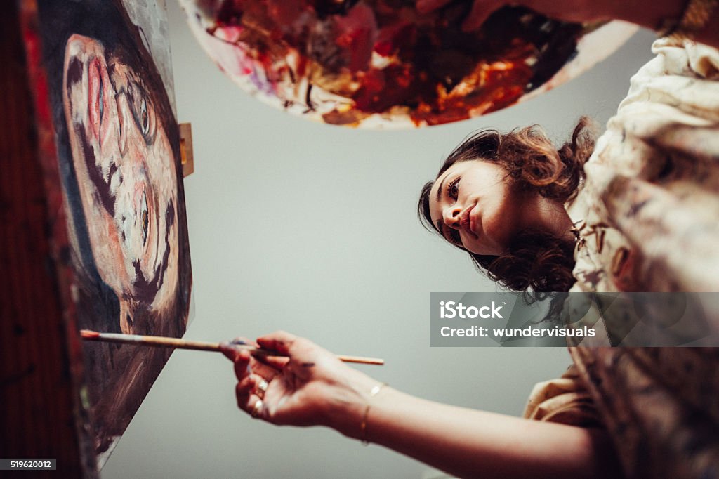 Artist painting on a canvas with brush and palette Low angle shot of a woman artist holding a palette while painting a canvas with a fine brush and wearing an artist's smock Painting - Activity Stock Photo