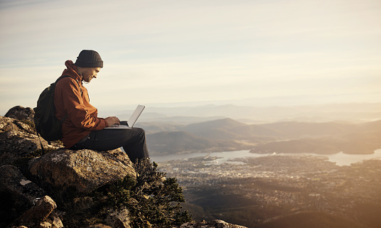 Shot of a young hiker using his laptop while sitting on top of a mountain