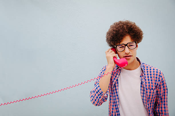 Portrait of young man talking on vintage telephone Serious young funny man talking on the pink telephone, isolated on blue wall background nerd teenager stock pictures, royalty-free photos & images