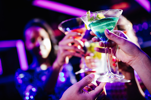 Close up of young adult hands holding colored cocktails and toasting in a night club