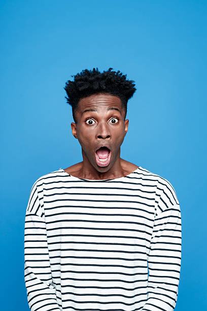 Shock, Afro american staring at camera with mouth open Studio portrait of afro american young man wearing striped top staring at the camera with mouth open. Studio portrait, blue background. gasping stock pictures, royalty-free photos & images