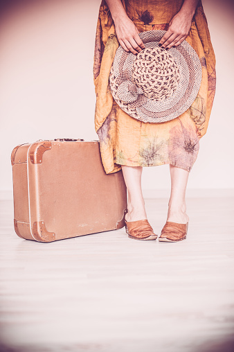 Woman standing with her vintage suitcases inside a home concept symbolizing she is going away on a trip or going on a vacation. Woman is wearing a brown patterned summer dress . Only showing the woman's legs, arms and shoes.Vertical sepia toned image. Copy space.  