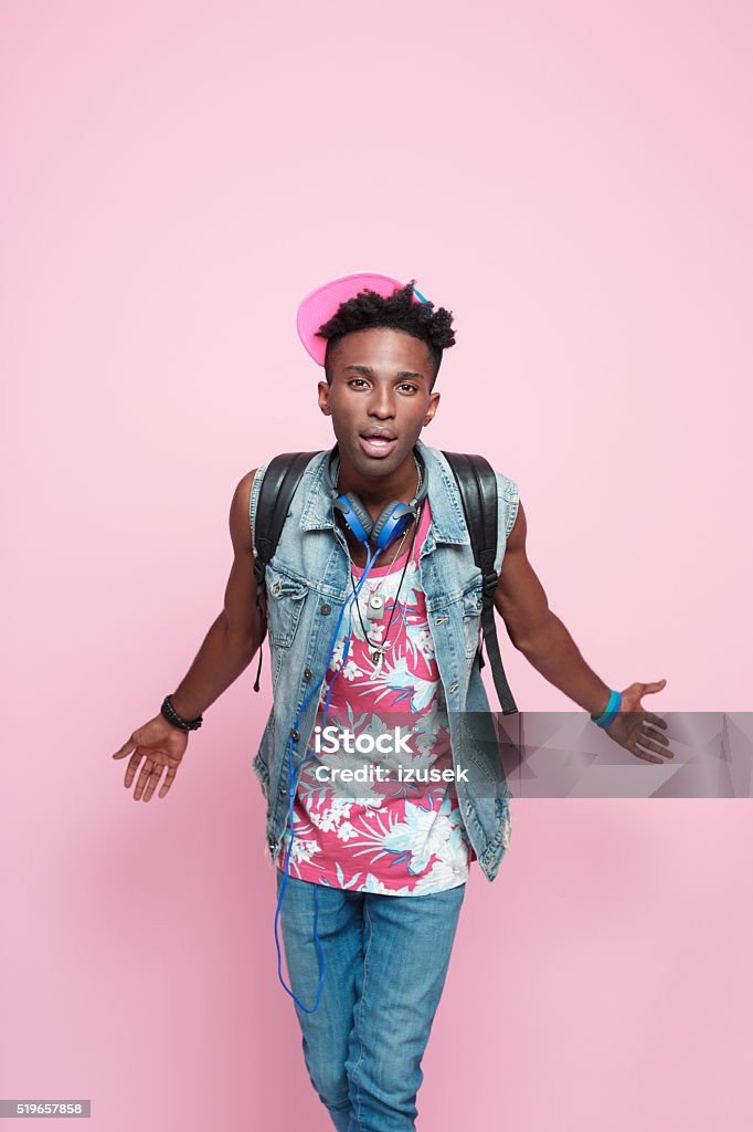 Cool afro american guy, summer studio portrait, pink background Summer portrait of carefree afro american young man wearing headphone, cap and jeans sleeveless jacket, standing against pink background and looking at the camera.  Adult Stock Photo