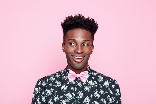 Portrait of surprised, carefree afro american young man wearing floral patern shirt and pink bow tie, looking away and smiling. Studio shot, pink background.