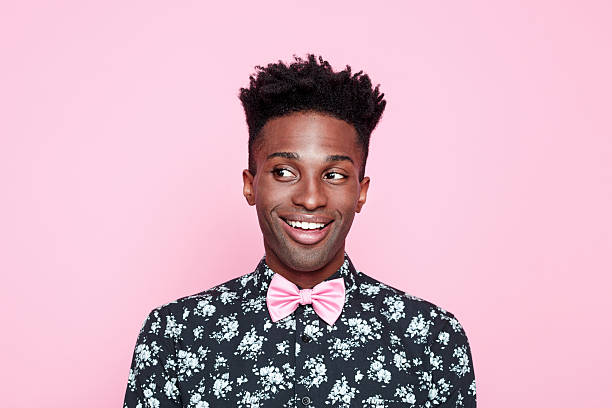 Funky afro american guy against pink background Portrait of surprised, carefree afro american young man wearing floral patern shirt and pink bow tie, looking away and smiling. Studio shot, pink background. fashion designer photos stock pictures, royalty-free photos & images
