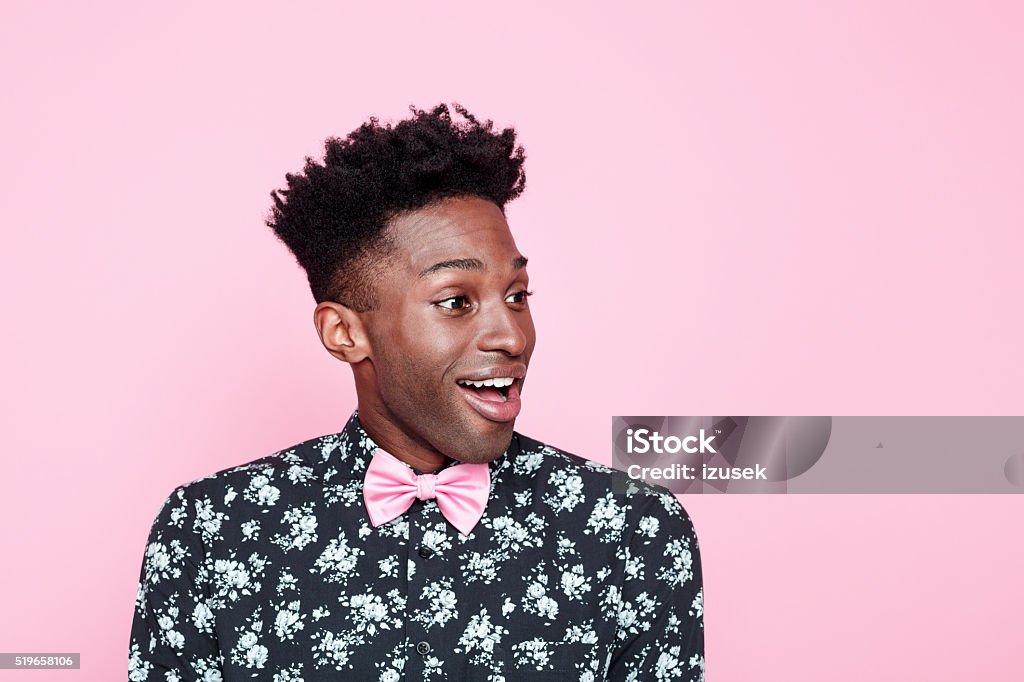 Excited afro american guy against pink background Portrait of excited, carefree afro american young man wearing floral patern shirt and pink bow tie, looking away and smiling. Studio shot, pink background. Men Stock Photo