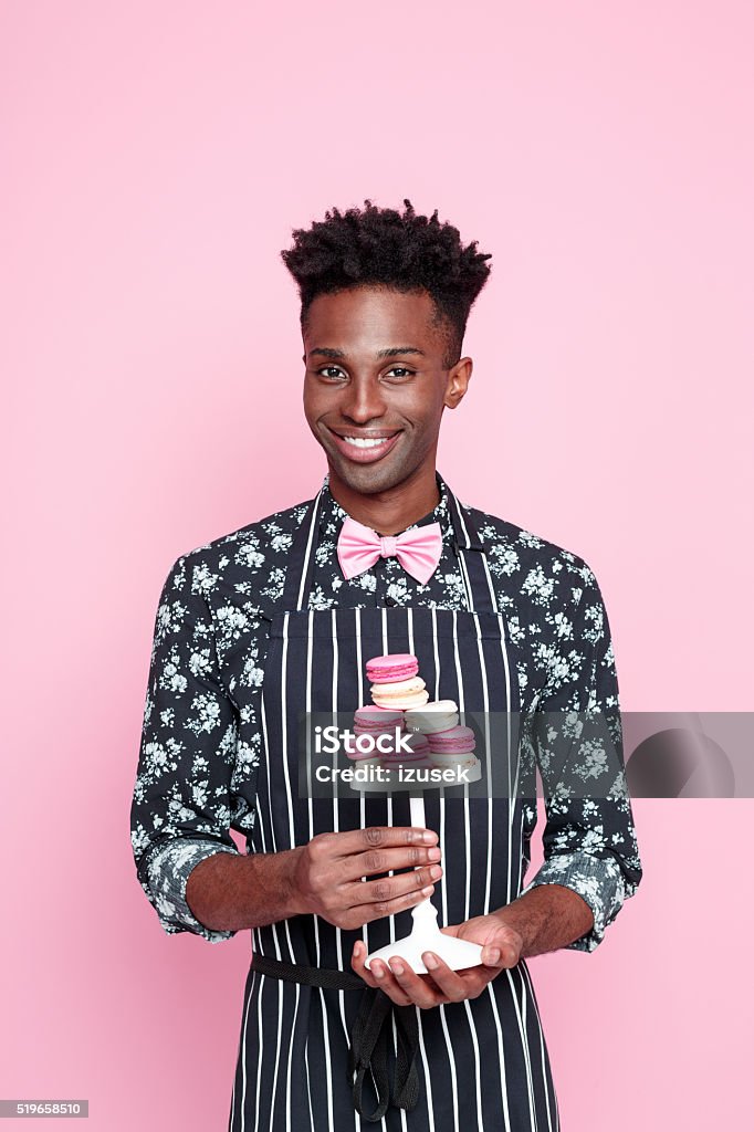 Cute afro american small business owner holding coockies Portrait of happy afro american young man wearing floral patern shirt, pink bow tie and apron, holding coockies, smiling at camera. Studio shot, pink background. Colored Background Stock Photo