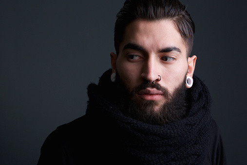 Close up portrait of a edgy modern young man with beard and piercings posing on gray background