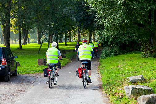 Essen, Germany - October 1, 2014: A group of caucasian senior men is cycling along promenade of river Ruhr in early autumn at warm and sunny day. Men are wearing yellow security vests and helmets. On vests of men in foreground are different  logos like Caritas and BMW. Scene is in Essen Werden.
