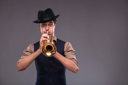 Waist-up portrait of a young handsome Caucasian jazz man in a suit with a black hat looking at the camera while holding a trumpet in his hand and playing on it, isolated on grey background with copy place