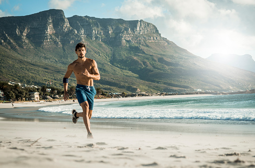 A picture of a very fit man running and exercising beneath a clear sky, along a beutiful beach and amazing mountains in the background. He is wearing just a blue shorts and black running sneakers. In the distance is the sea and the sun is giving a nice warm light