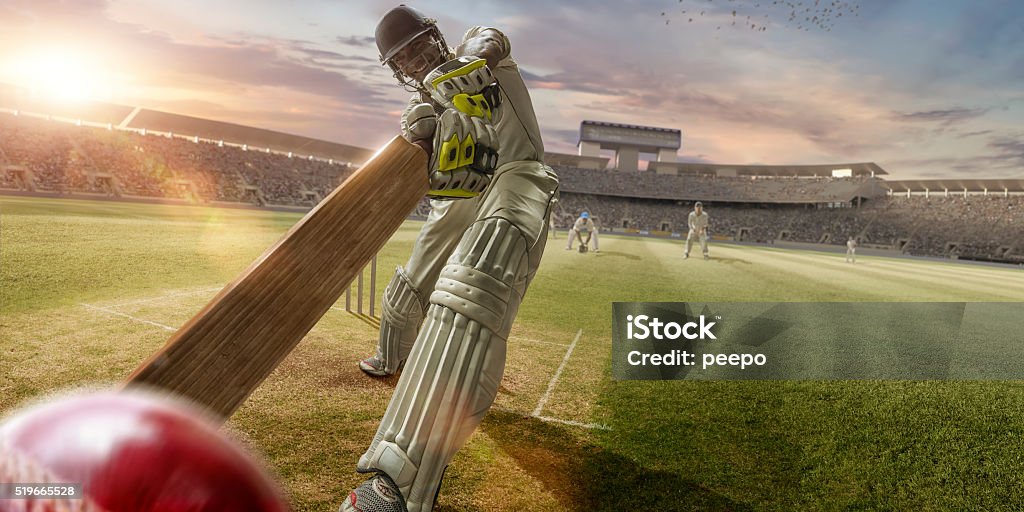 Cricket Batsman Hitting Ball During Cricket Match In Stadium A close up image of a professional cricketer playing in batsman position wearing cricket whites and safety helmet, having just hit a ball during a cricket match in an outdoor stadium full of spectators. The action occurs under an evening sky at sunset. Stadium is fake, created from photographic and CG elements.  Sport of Cricket Stock Photo