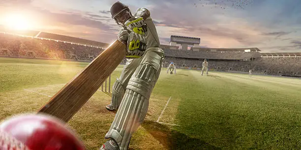 A close up image of a professional cricketer playing in batsman position wearing cricket whites and safety helmet, having just hit a ball during a cricket match in an outdoor stadium full of spectators. The action occurs under an evening sky at sunset. Stadium is fake, created from photographic and CG elements. 