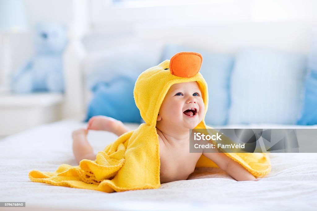 Funny cute baby after bath in yellow duck towel Happy laughing baby wearing yellow hooded duck towel sitting on parents bed after bath or shower. Clean dry child in bedroom. Bathing and washing of little kids. Children hygiene. Textile for infants. Baby - Human Age Stock Photo