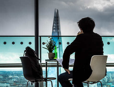 Rear view image of an unrecognisable male sitting at a table, looking out of a window over the sprawling urban landscape of the City of London, UK. His backpack is perched in the other seat, and a green bottle of beer is on the table. In the center of the frame, slightly defocused, is the tallest building in western Europe, The Shard. The man is drinking his beer from a glass, and his left hand is rested on his thigh. Colour image with lots of room for copy space.