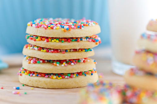 A stack of rainbow-sprinkled biscuits sit amongst cooking paraphernalia. Shallow depth-of-field, emphasis on blues, pinks, and yellows.