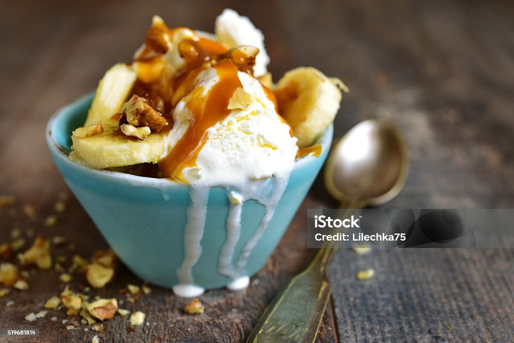 Vanilla ice cream with walnuts,banana and caramel. Vanilla ice cream with walnuts,banana and caramel in a blue bowl on a rustic wooden background. Banana Stock Photo