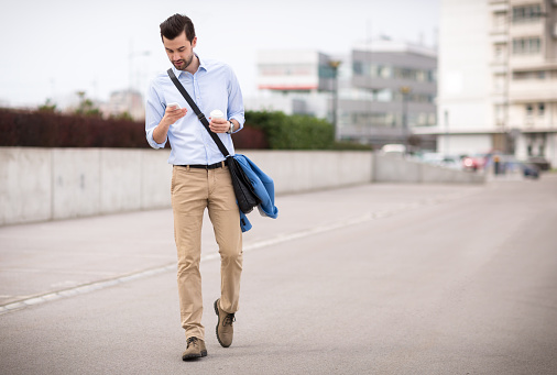 Young businessman is on the phone while drinking coffee. He is wearing white shirt, khaki pants and folded blue jacket. He is wearing beard and slicked hair. He has his bag over his shoulder. The photography taken with shallow depth of field.