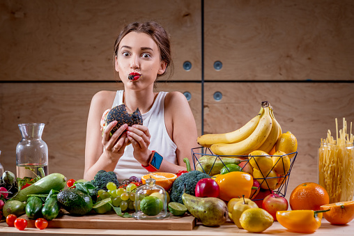 Hungry woman eating black burger at the table full of fruits and vegetables on the wooden background. Choosing between healthy and unhealthy food