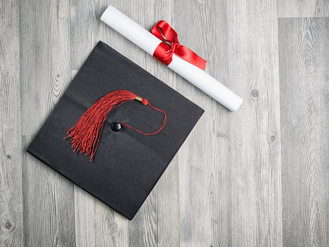 Close up view of black graduation cap on gray wooden background .The object was photographed from a high angle viewpoint.No people are seen in frame.The diploma scroll has a red ribbon.Shot with medium format camera Hasselblad in studio.