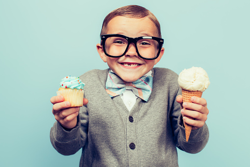 A young nerd boy with glasses loves eating sugary treats such as ice cream and cupcakes. He is smiling broadly and has frosting on his nose. The boy is wearing glasses and a bow tie. Retro styled  