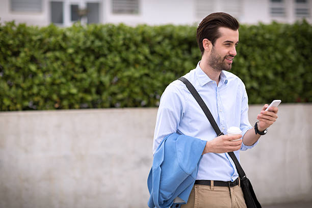 Businessman using modern technology Young businessman uses cell phone. He is wearing white shirt, khaki pants and folded blue jacket. He is wearing beard and slicked hair. He has his bag over his shoulder. The photography taken with shallow depth of field. slicked back hair stock pictures, royalty-free photos & images