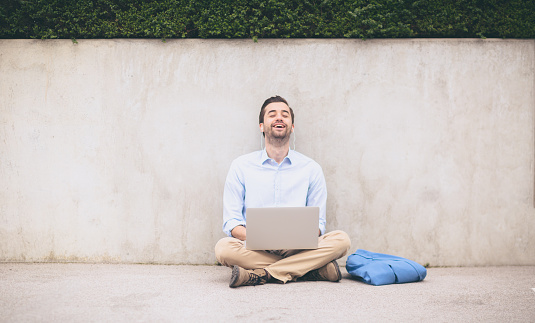 Young businessman is relaxing next to concrete wall. He is wearing white shirt, khaki pants and has folded blue jacket. He is wearing beard and slicked hair. The photography taken with shallow depth of field.